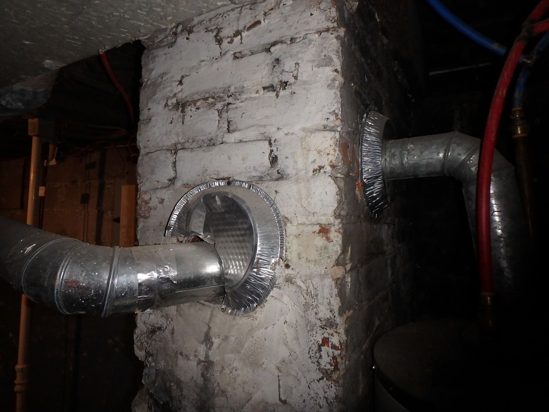 Ductwork attached to a weathered brick chimney. Silver ducting with silver tape fittings. Dark basement setting.