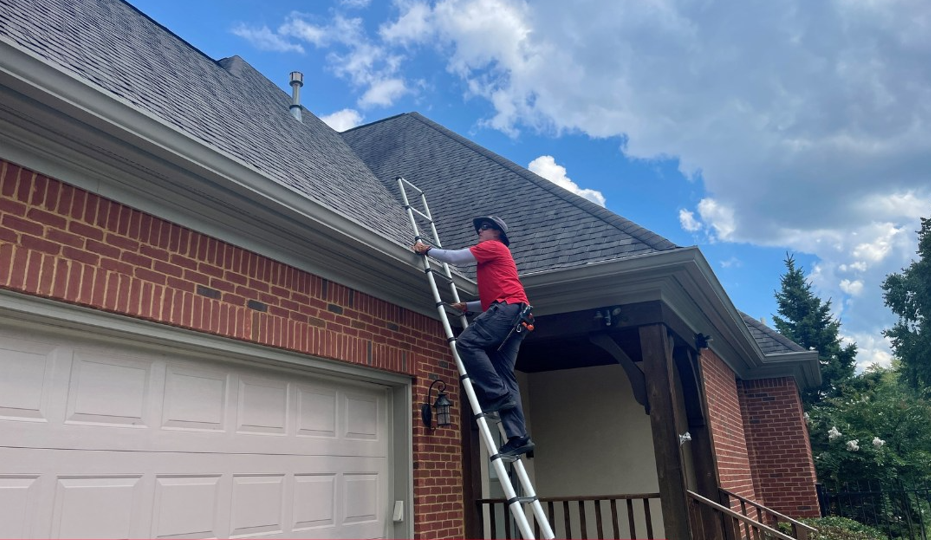 Person on a ladder cleaning a house gutter on a sunny day; red shirt, brick house.