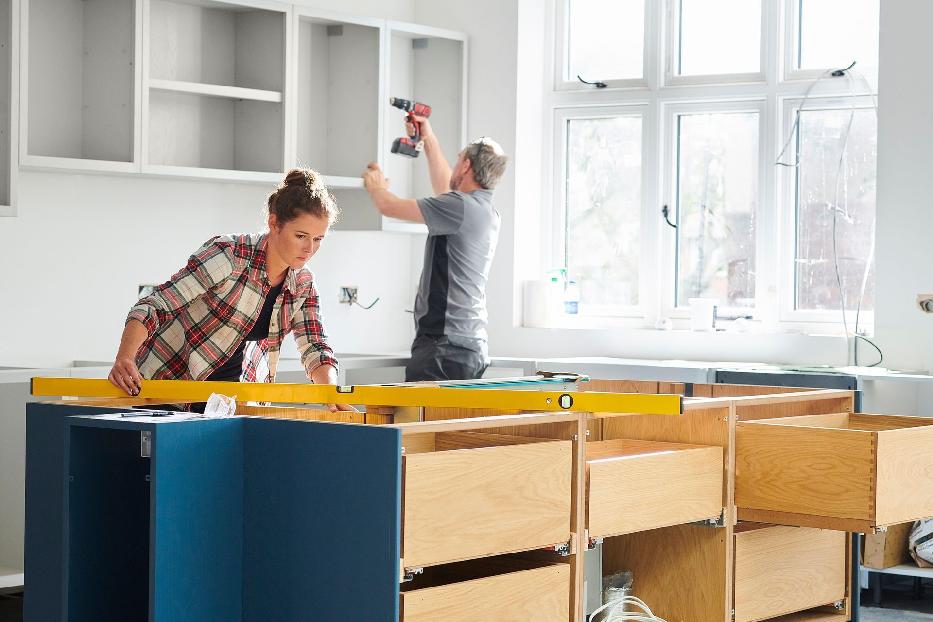Two people installing kitchen cabinets with tools, working near a window.
