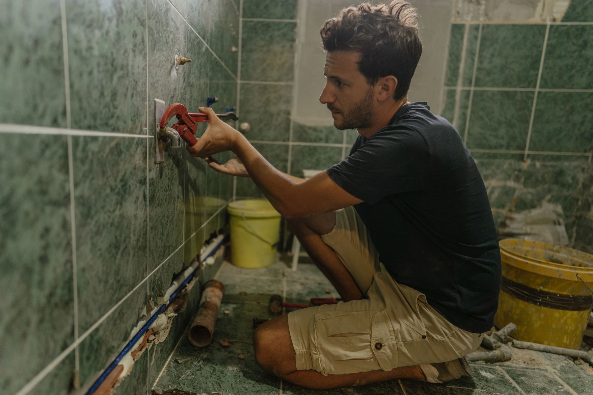 Man using a wrench to work on plumbing in a bathroom with green tiles; crouched on floor.