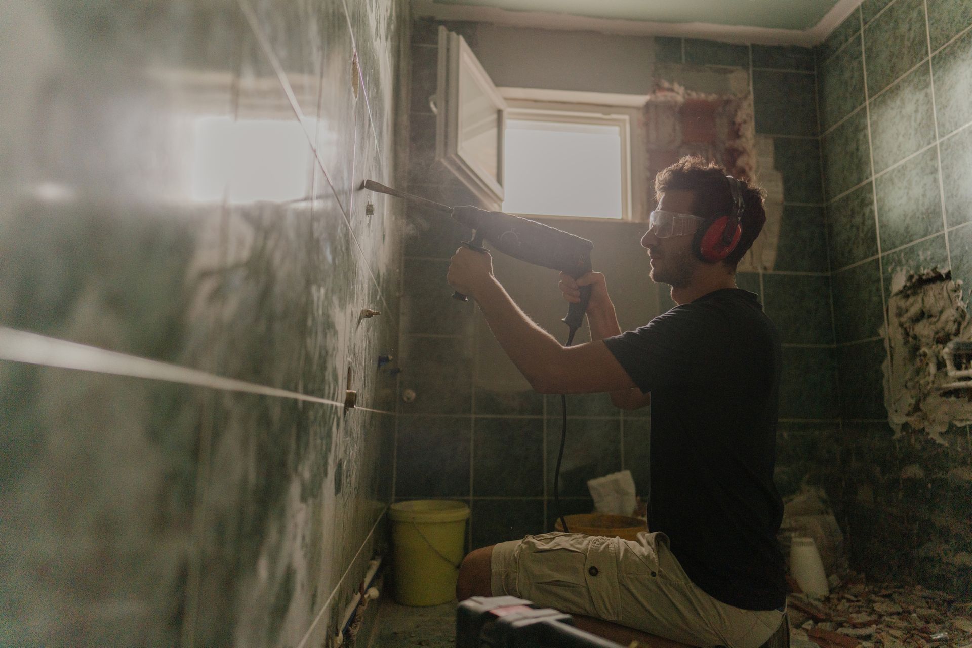 Person drills into tiled bathroom wall, wearing ear protection and safety glasses.