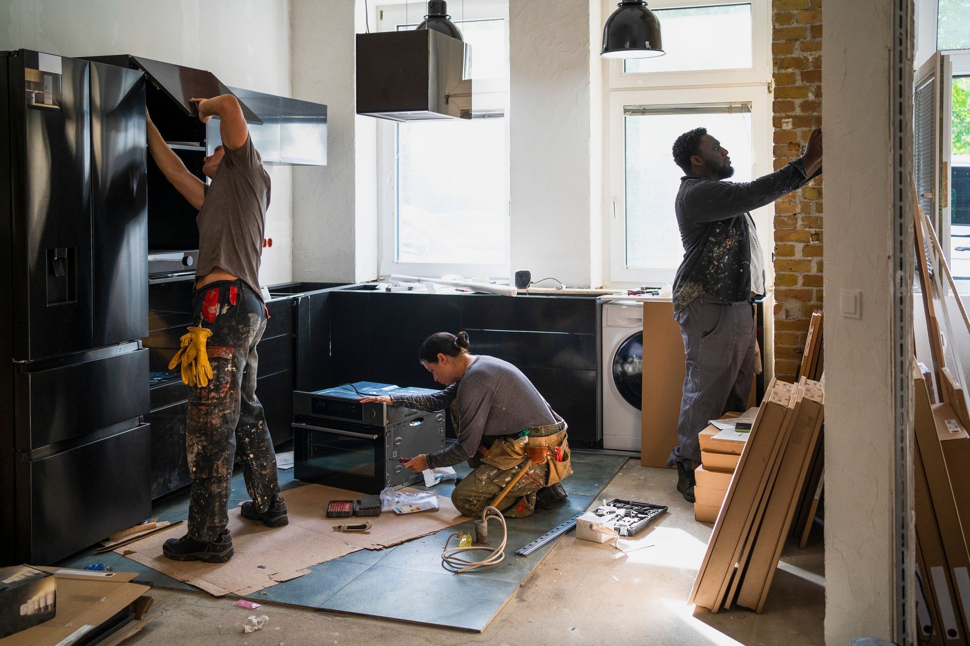 Three workers installing a black kitchen: one on refrigerator, one on oven, one on wall. Indoors.