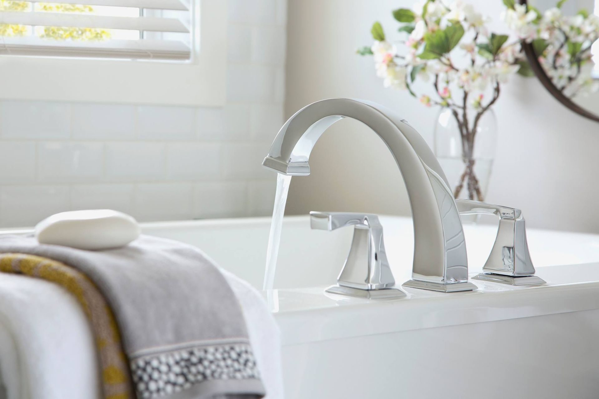 Bathtub faucet with water running. Towel and soap on tub rim, flowers in background.