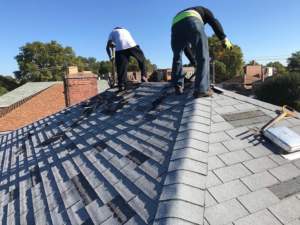 Two people repairing a shingled roof under a blue sky, using tools.
