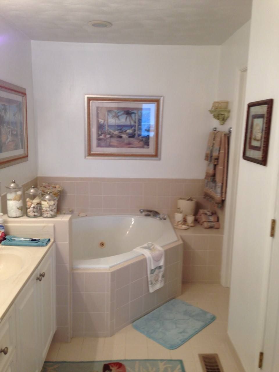 Bathroom with a corner jacuzzi tub, light tile, and artwork. A blue bath mat is on the floor.