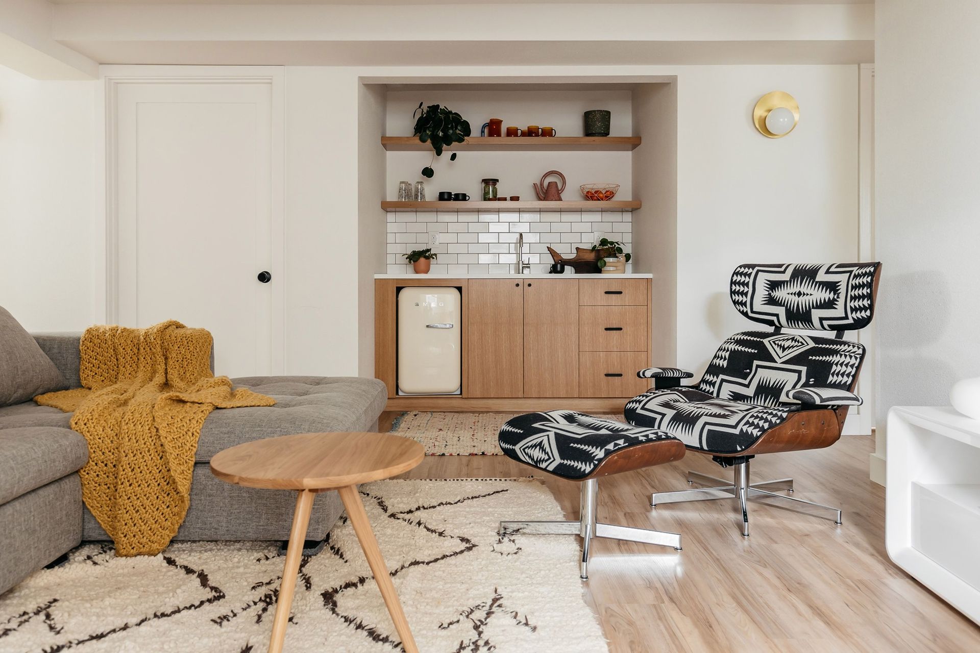 Living room with built-in bar: gray sofa, patterned chair, wooden table, mini-fridge, and shelves with décor.