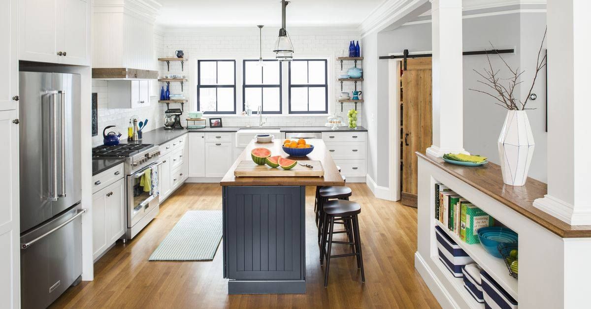 Modern white kitchen with stainless steel appliances, dark blue island, wood accents, and three windows.