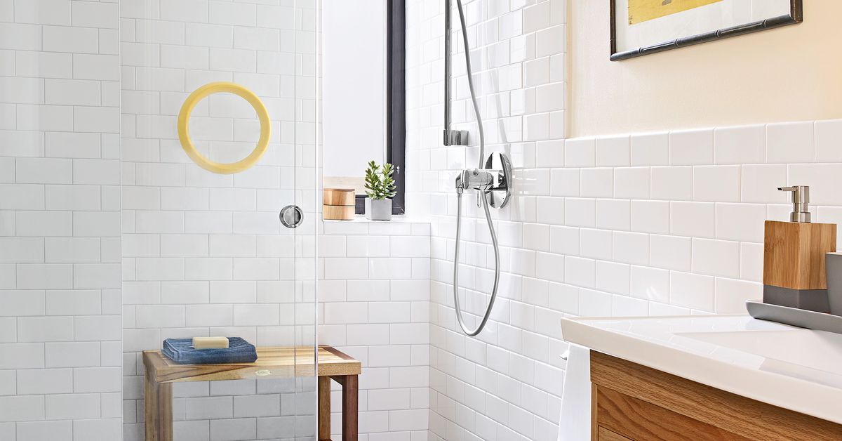 White-tiled bathroom with wooden vanity, showerhead, and bench; a yellow-framed mirror and small window are visible.
