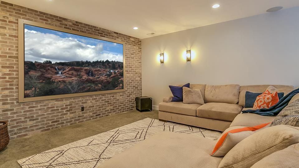 Home theater with large screen on brick wall, beige sectional, rug, sconces, and overhead lighting.