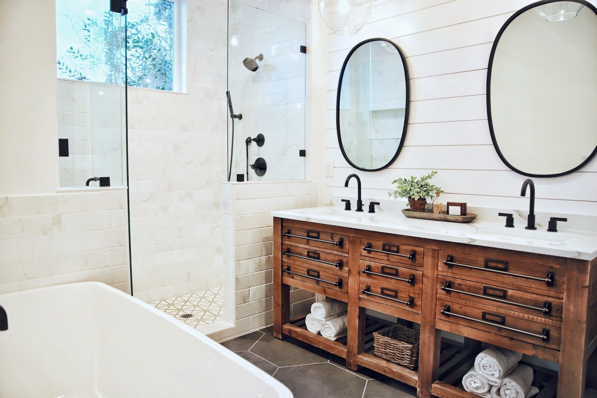 Bathroom with a wooden vanity, oval mirrors, and a glass shower. White walls and marble countertop.