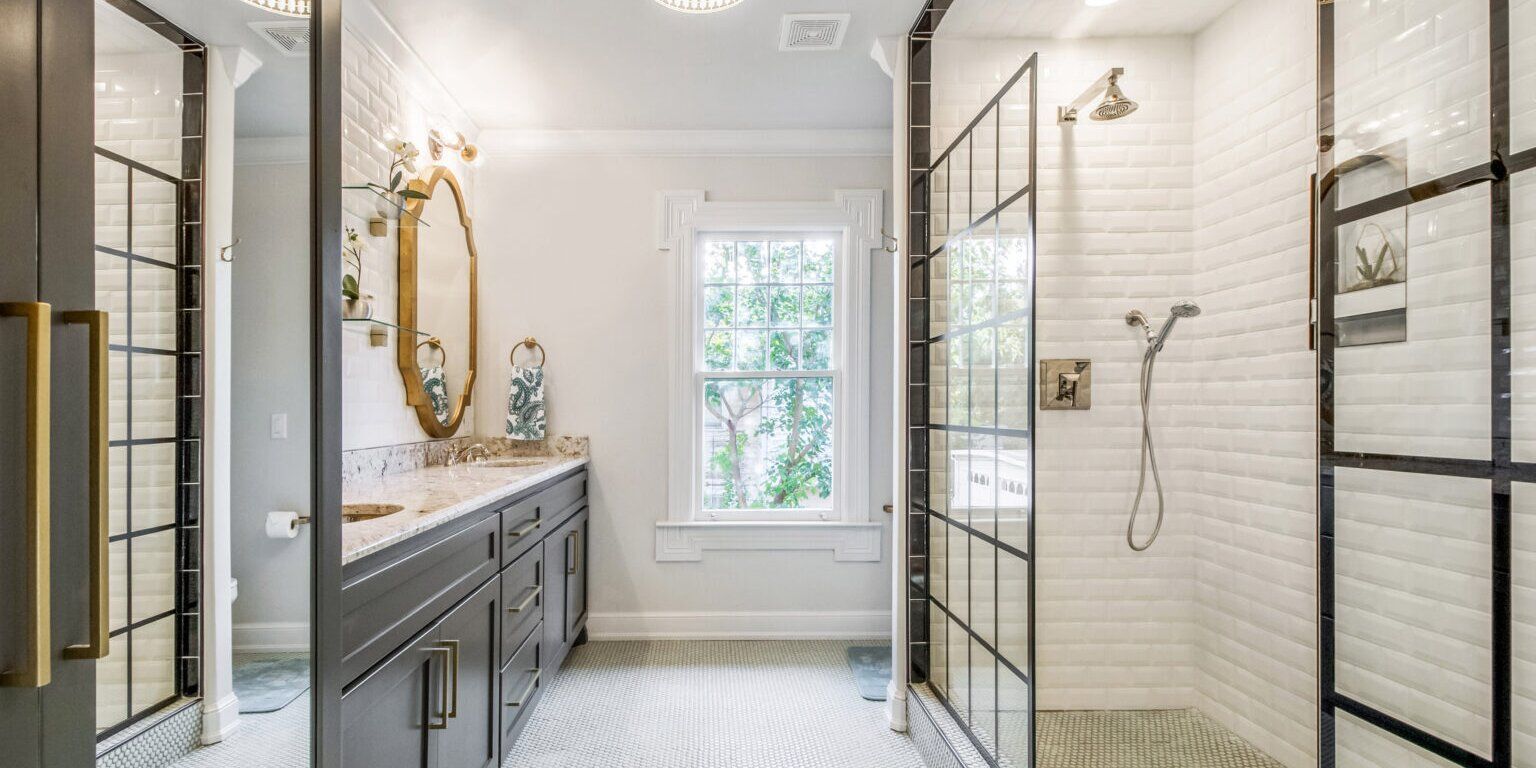Bathroom with shower behind black-framed glass doors, vanity with gold mirror, and white brick tiles.