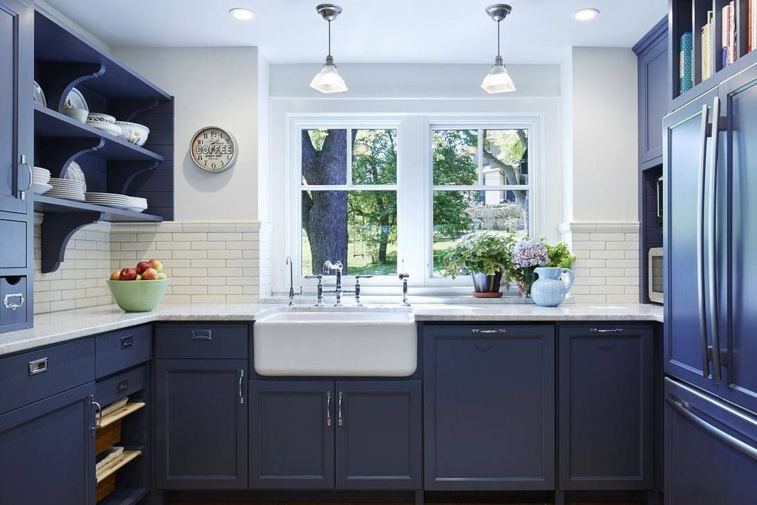 Navy blue kitchen with farmhouse sink, window, white backsplash, and overhead lights.