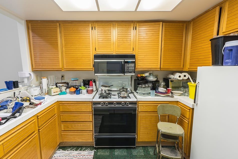 A cluttered kitchen with light wood cabinets, stove, microwave, and a small chair.