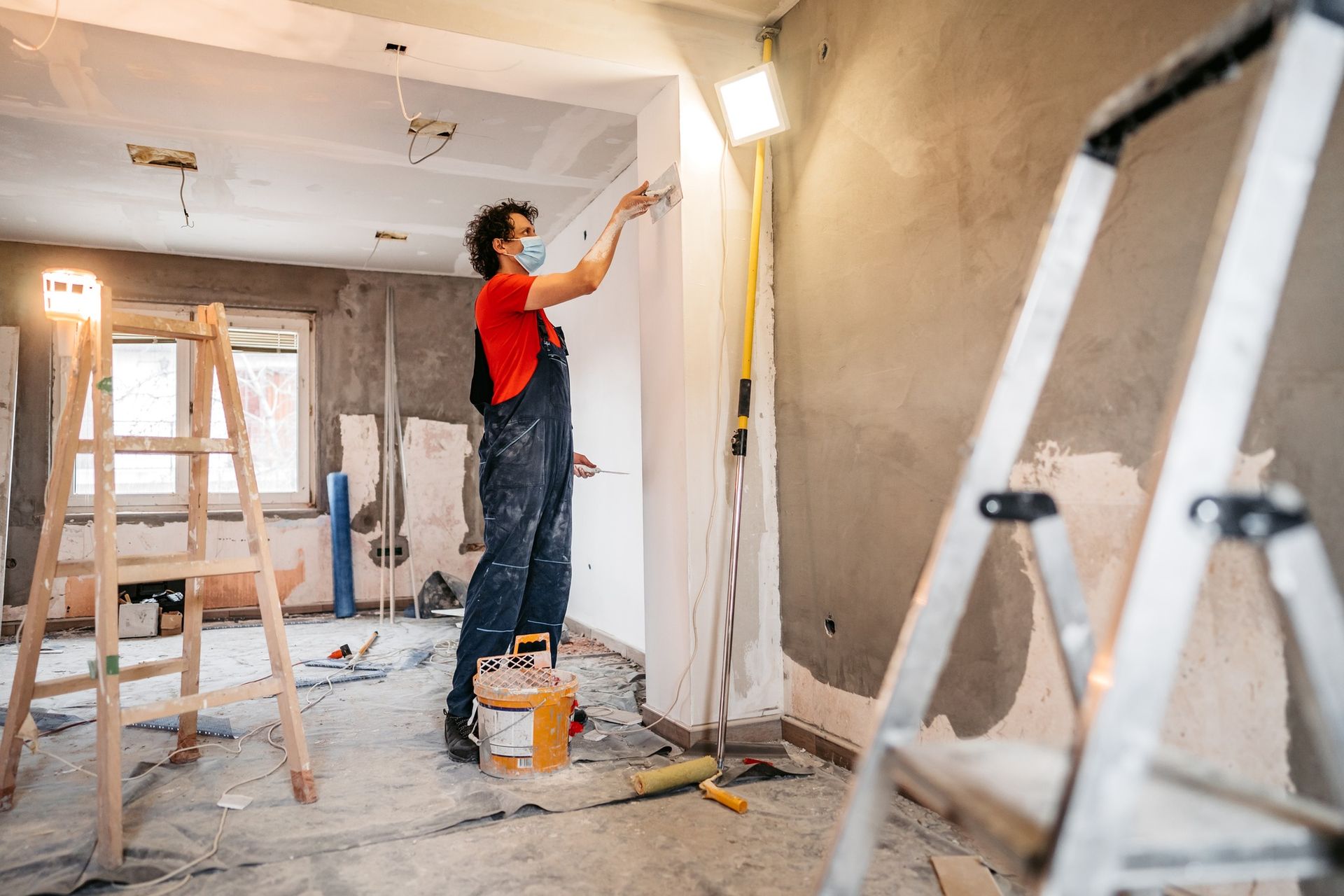 Person in overalls paints a wall in a room under renovation, with ladders and tools visible.
