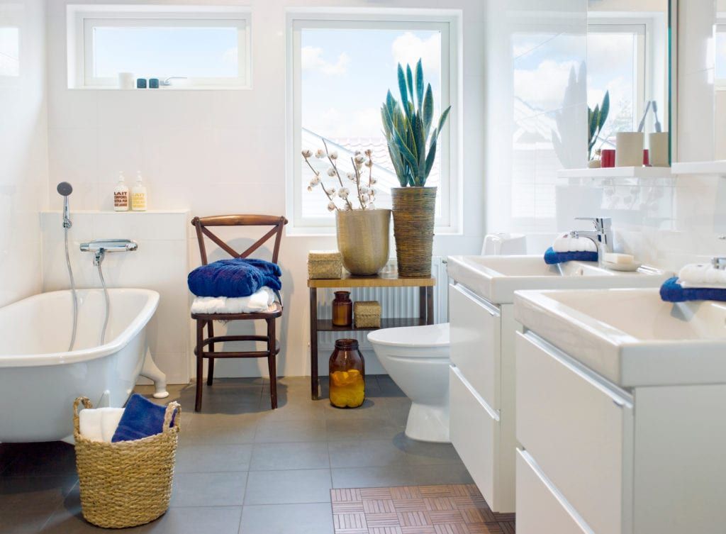 Bright white bathroom with a tub, two sinks, and a large window with plants.