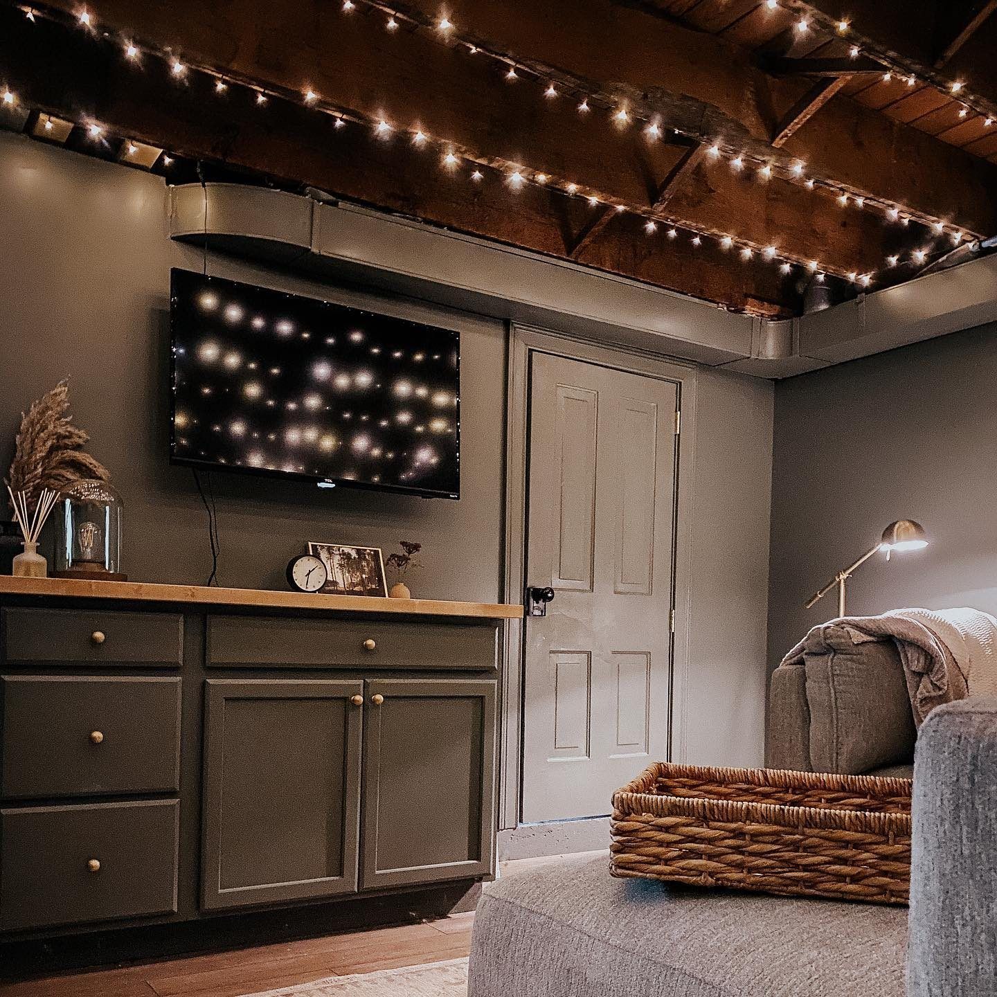 Cozy basement room with TV, string lights, and gray cabinets. Wicker basket on couch.