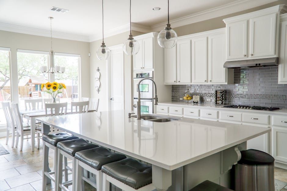 White kitchen with a large island, cabinets, and a dining area, featuring a sink and a row of stools.
