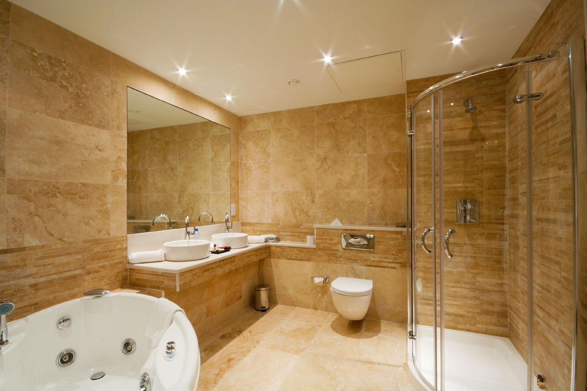 Luxurious bathroom with tan stone walls and flooring. Features a jacuzzi tub, shower, toilet, and double sinks.