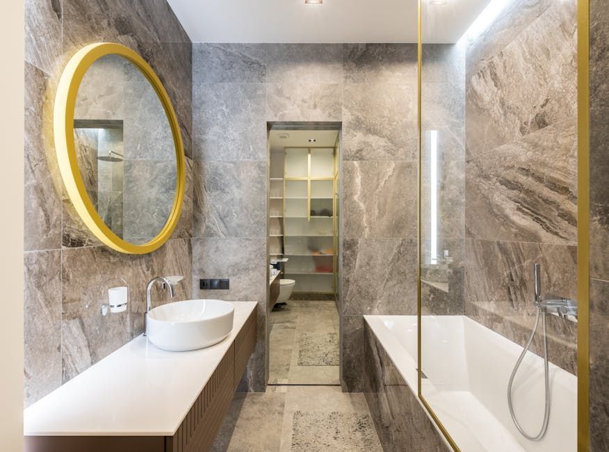 Modern bathroom with marble-like walls, gold-framed mirror, white sink, bathtub, and a view into a closet.