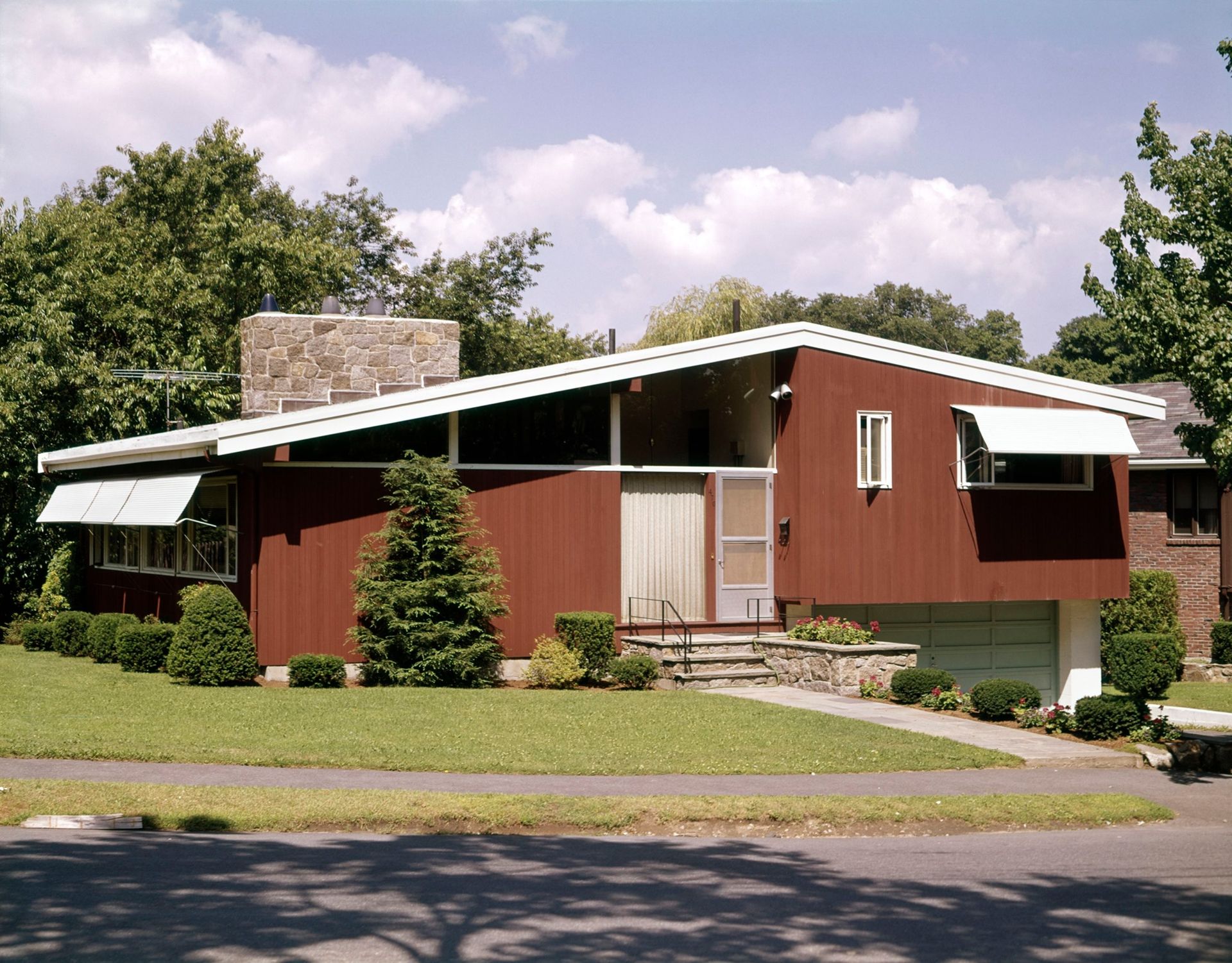 Mid-century modern home with brown siding, white roof, and awnings; green lawn and trees.