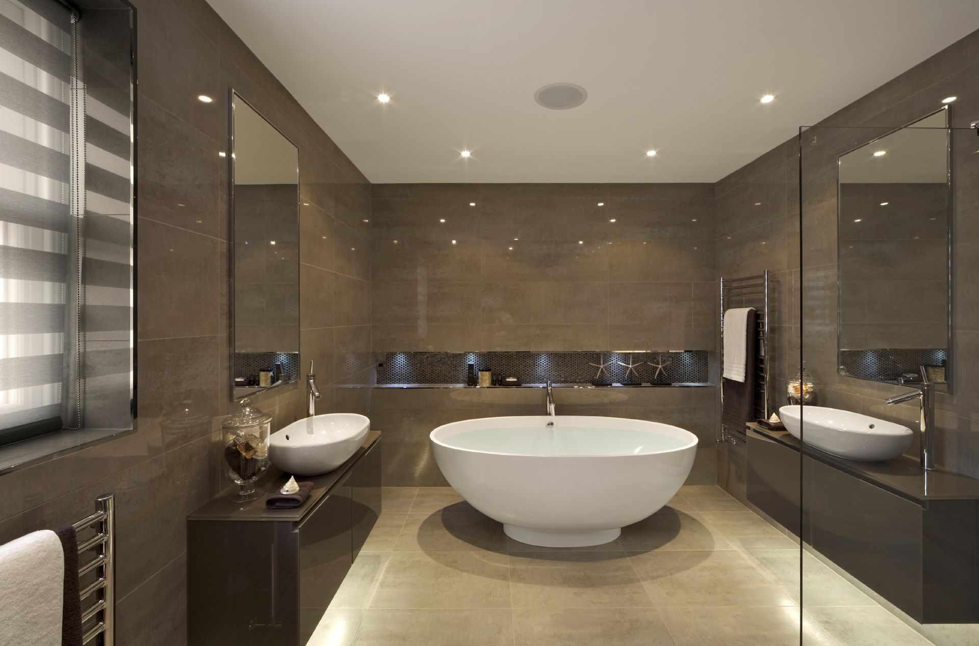 Modern bathroom with a white tub, two sinks, mirrors, and brown tile walls.