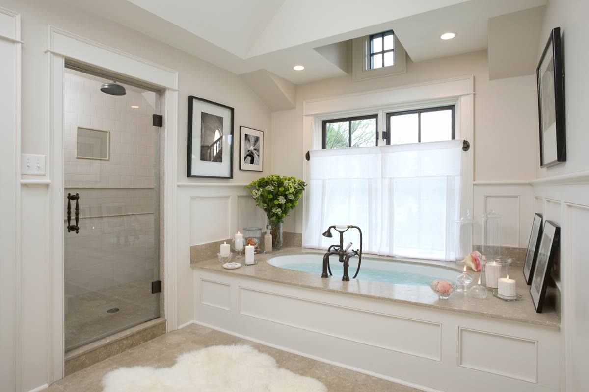 Bathroom with a built-in tub, glass shower, and white walls. Light tan countertop and a small window with sheer curtains.