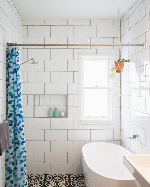 White-tiled bathroom with shower, window, and freestanding tub. Blue patterned shower curtain and potted plant.