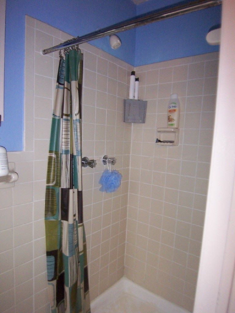 Shower stall with blue ceiling and beige tile. Shower curtain with colorful squares. Soap and shampoo on wall.