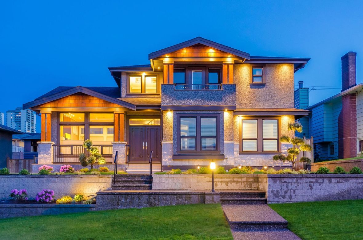 Two-story house with illuminated porch and windows at dusk. Brick and stone facade, pathway to front door.