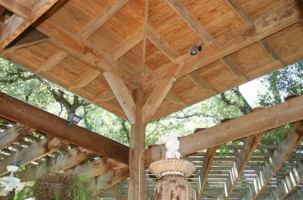 Wooden gazebo ceiling with beams and light, viewed from below with trees in the background.
