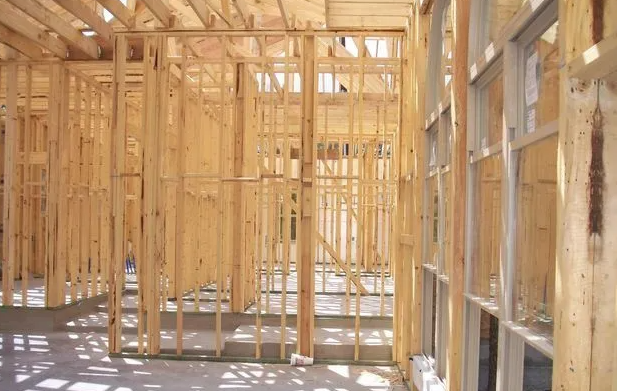 Interior view of a building under construction, showing wooden framing, windows, and unfinished floor.