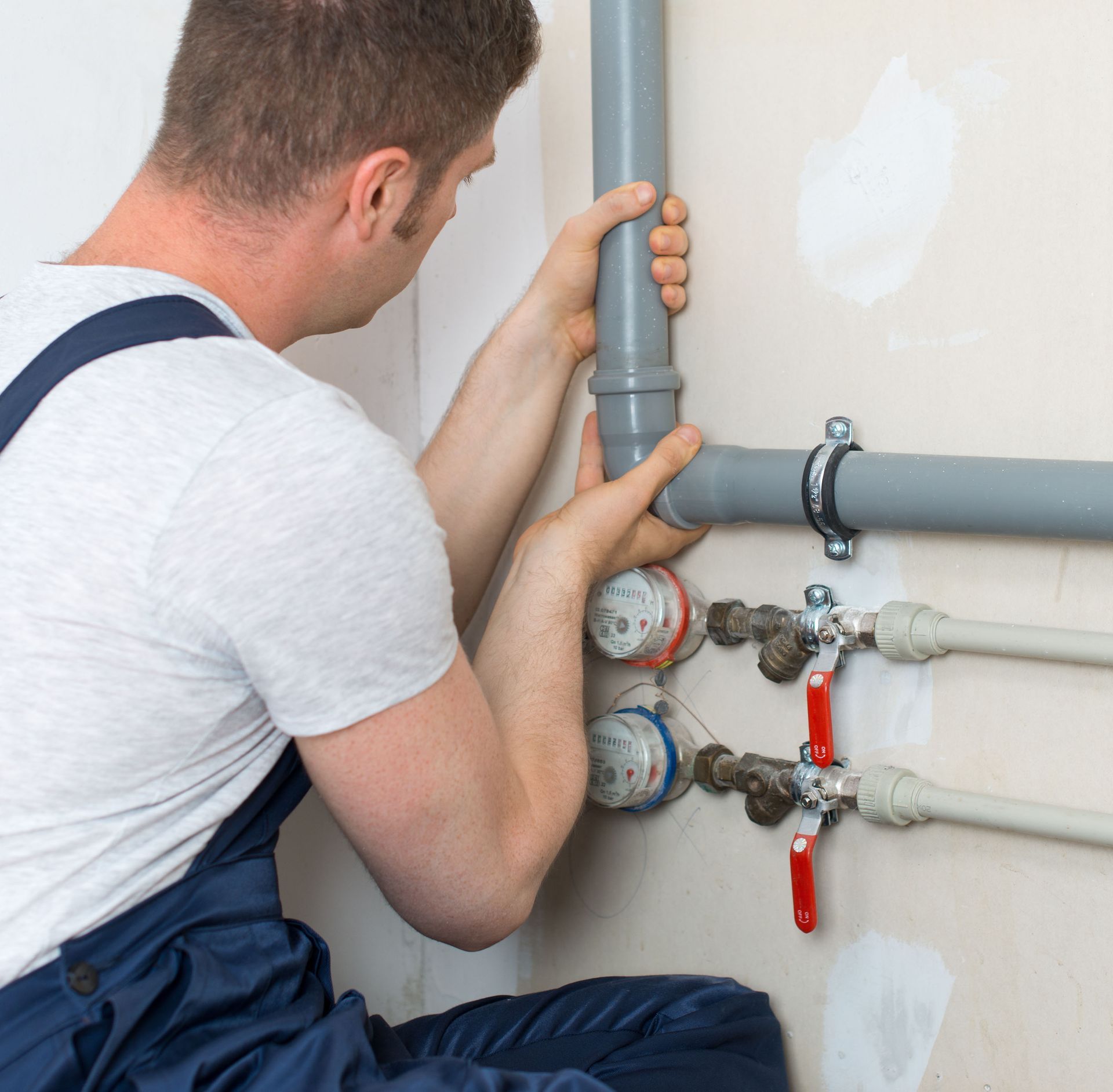 Plumber in blue overalls connecting gray pipes near water shut-off valves on a beige wall.
