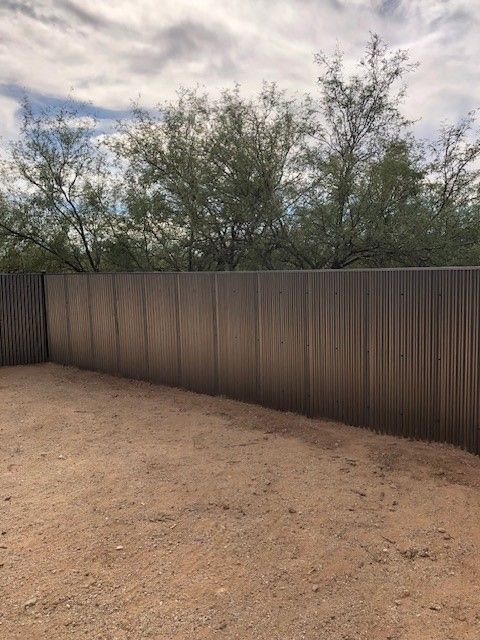 a brown corrugated fence surrounds a dirt field with trees in the background
