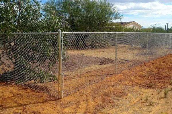 A chain link fence surrounds a dirt field with trees in the background.