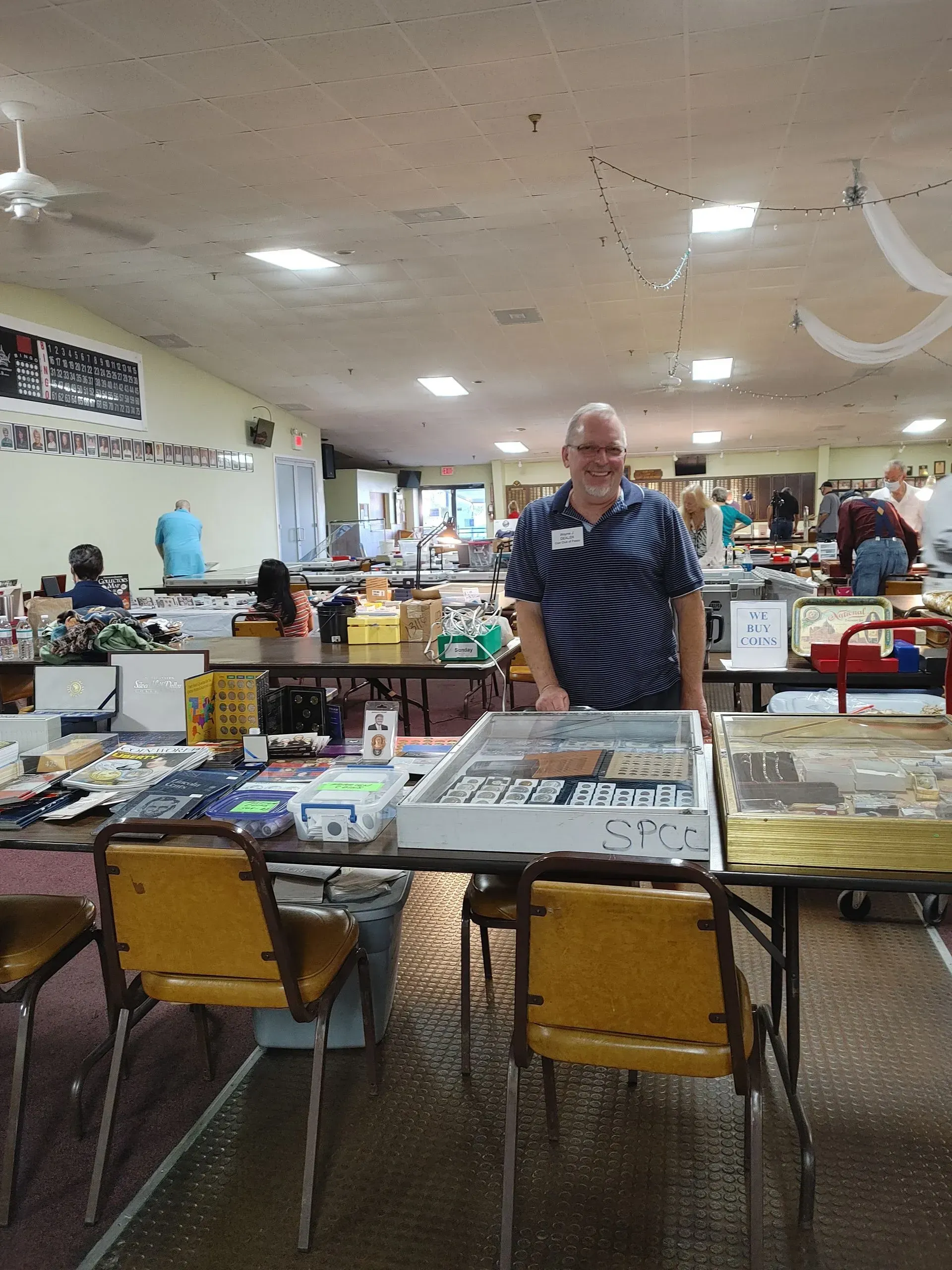 Man at a table with items for sale at an indoor event. He smiles at the camera.