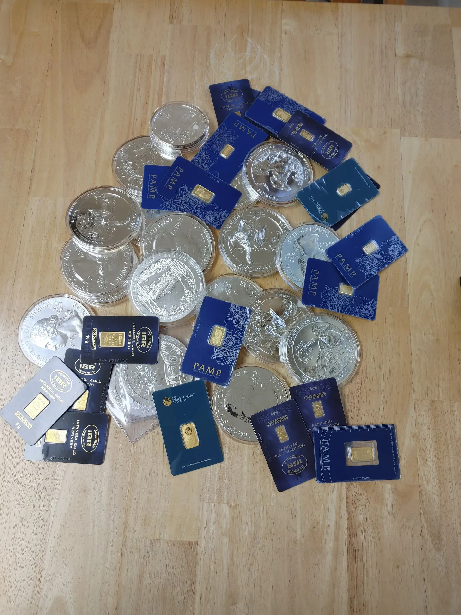 Pile of gold bars in blue cases and silver coins on a wooden surface.