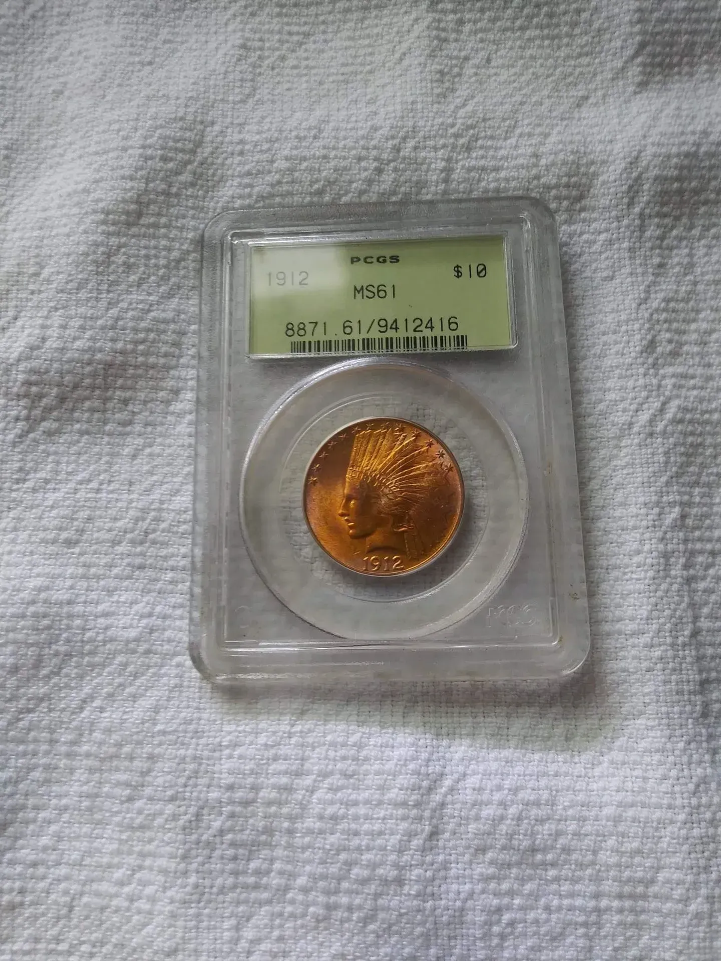 A gold-colored coin in a clear plastic case, against a white fabric background. The case has a grading label.