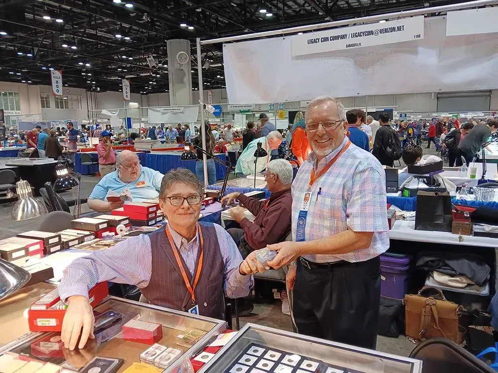 Two people at a coin show booth, one showing coins. Many people and tables with coins are in the background.