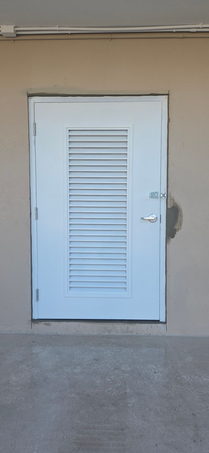 White door with horizontal vents in a tan building. Concrete floor below.
