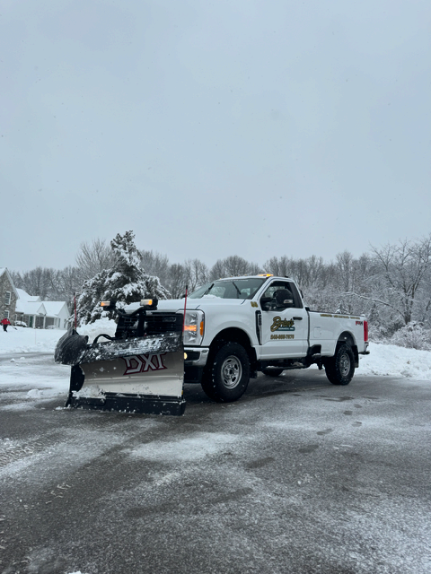 A white truck with a snow plow attached to it is parked in a parking lot.