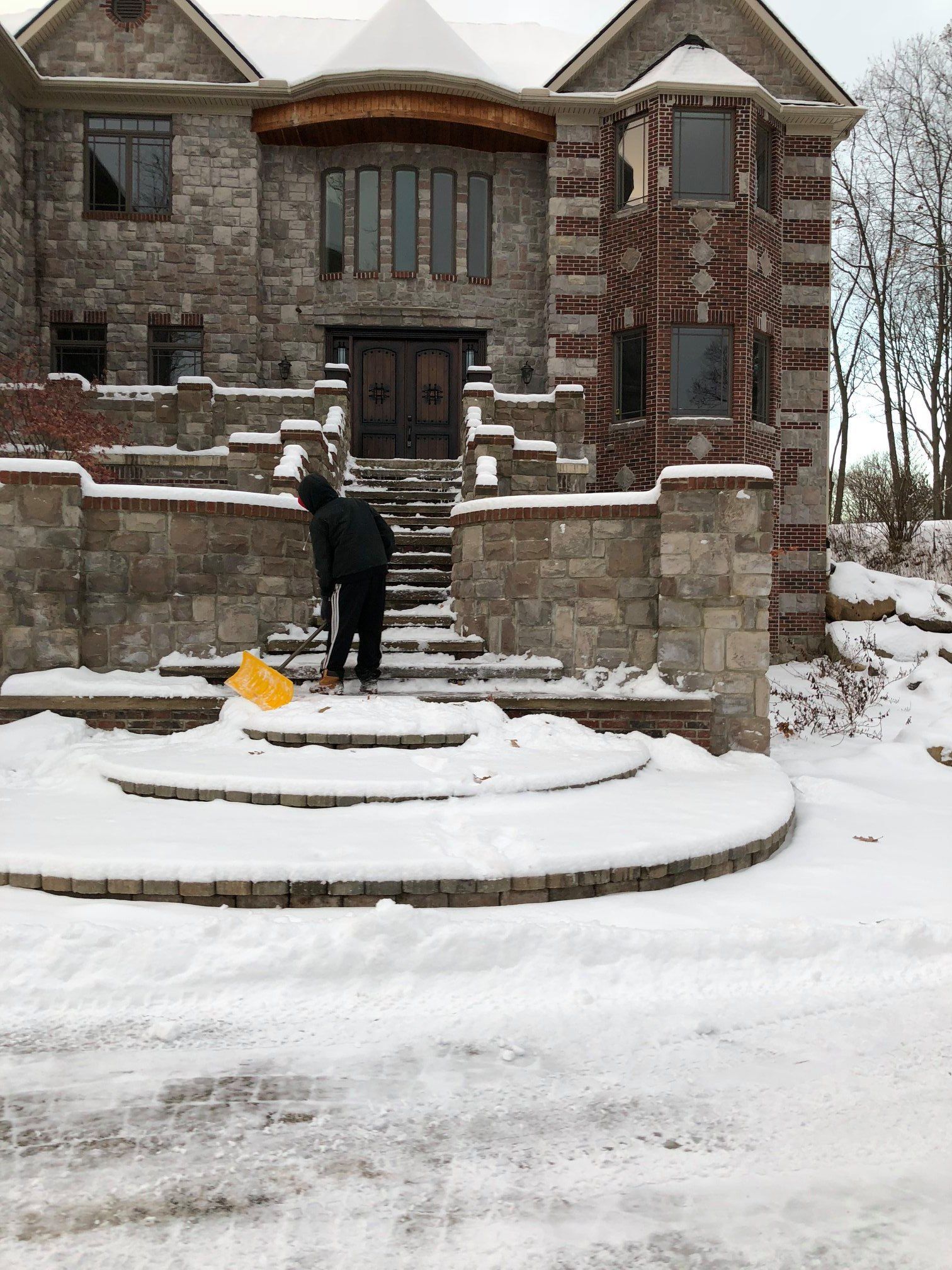 A man is shoveling snow in front of a large brick house.