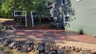 Brick patio next to a house with a deck and a rock retaining wall.