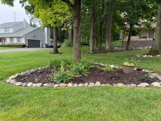 A garden bed with brown mulch and river rocks surrounding plants next to trees and grass.