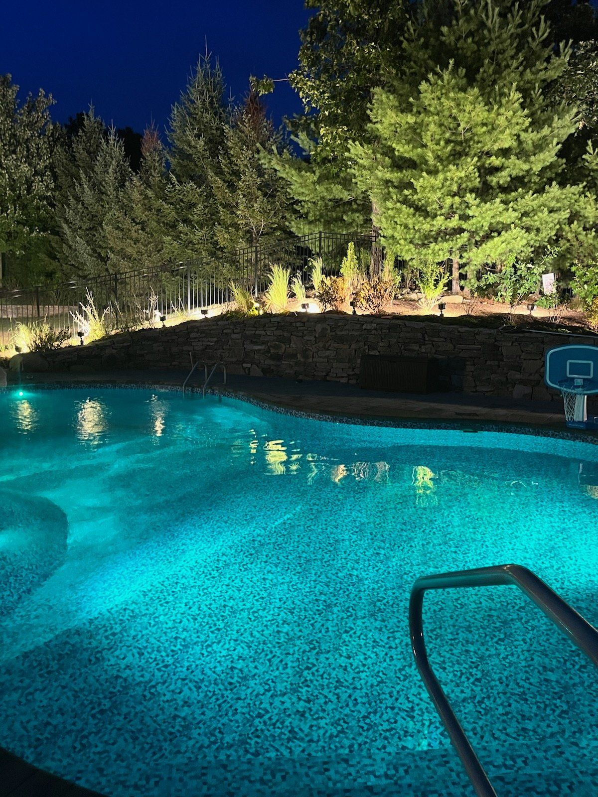 A swimming pool is lit up at night with a basketball hoop in the background.