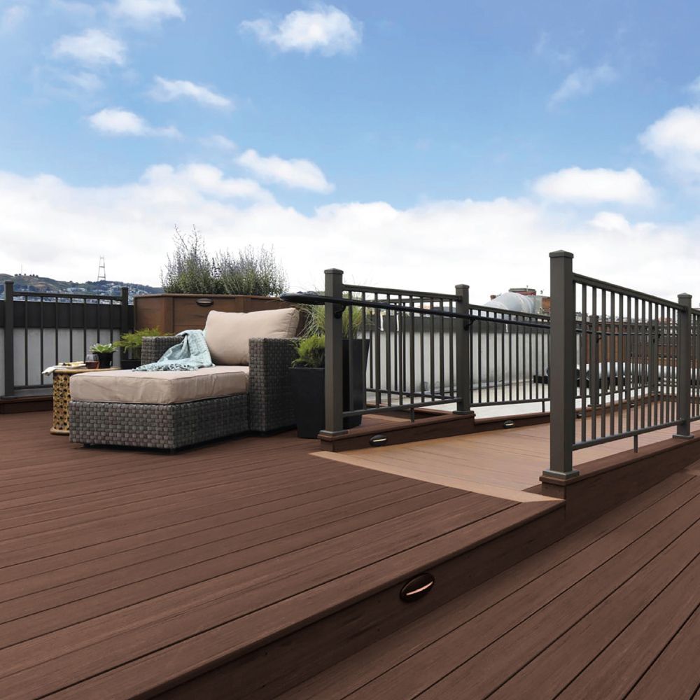 A rooftop deck with brown composite decking, gray furniture, and black railing overlooks a cityscape under a blue sky.