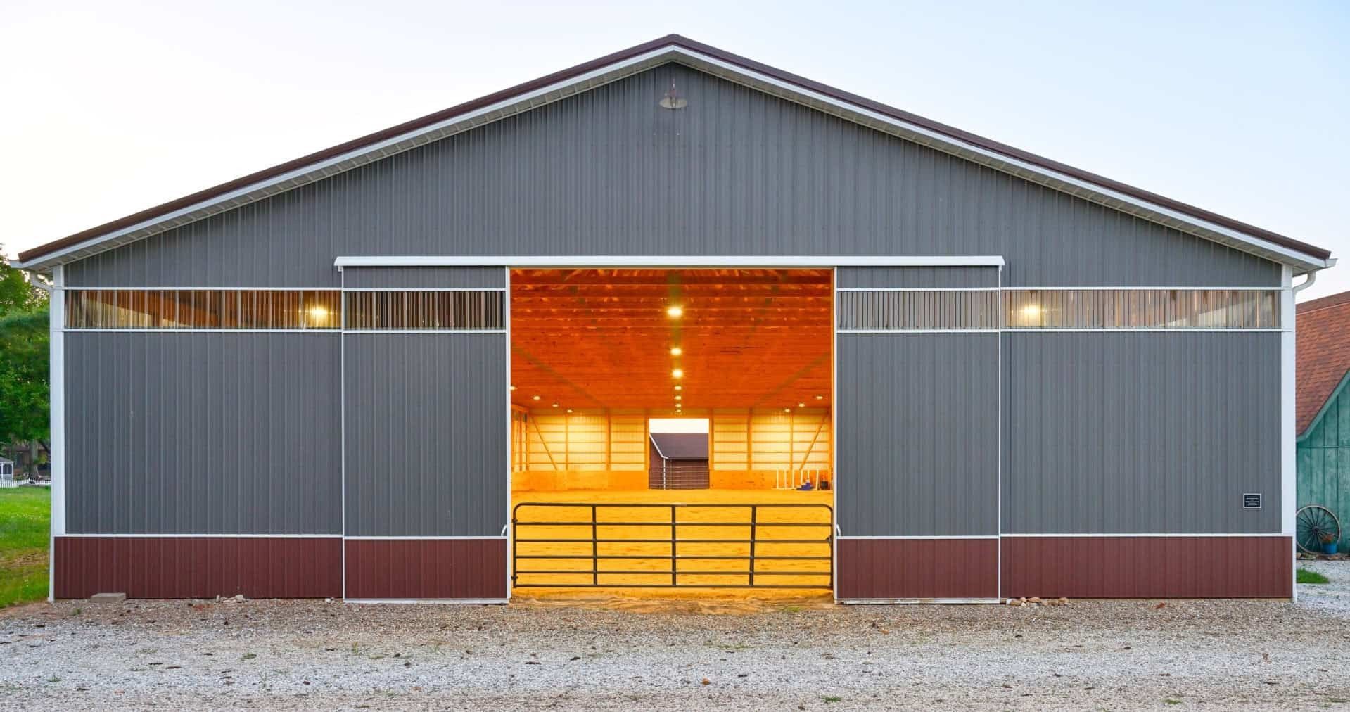 Front view of a gray metal barn with a lit central entrance and gravel foreground.