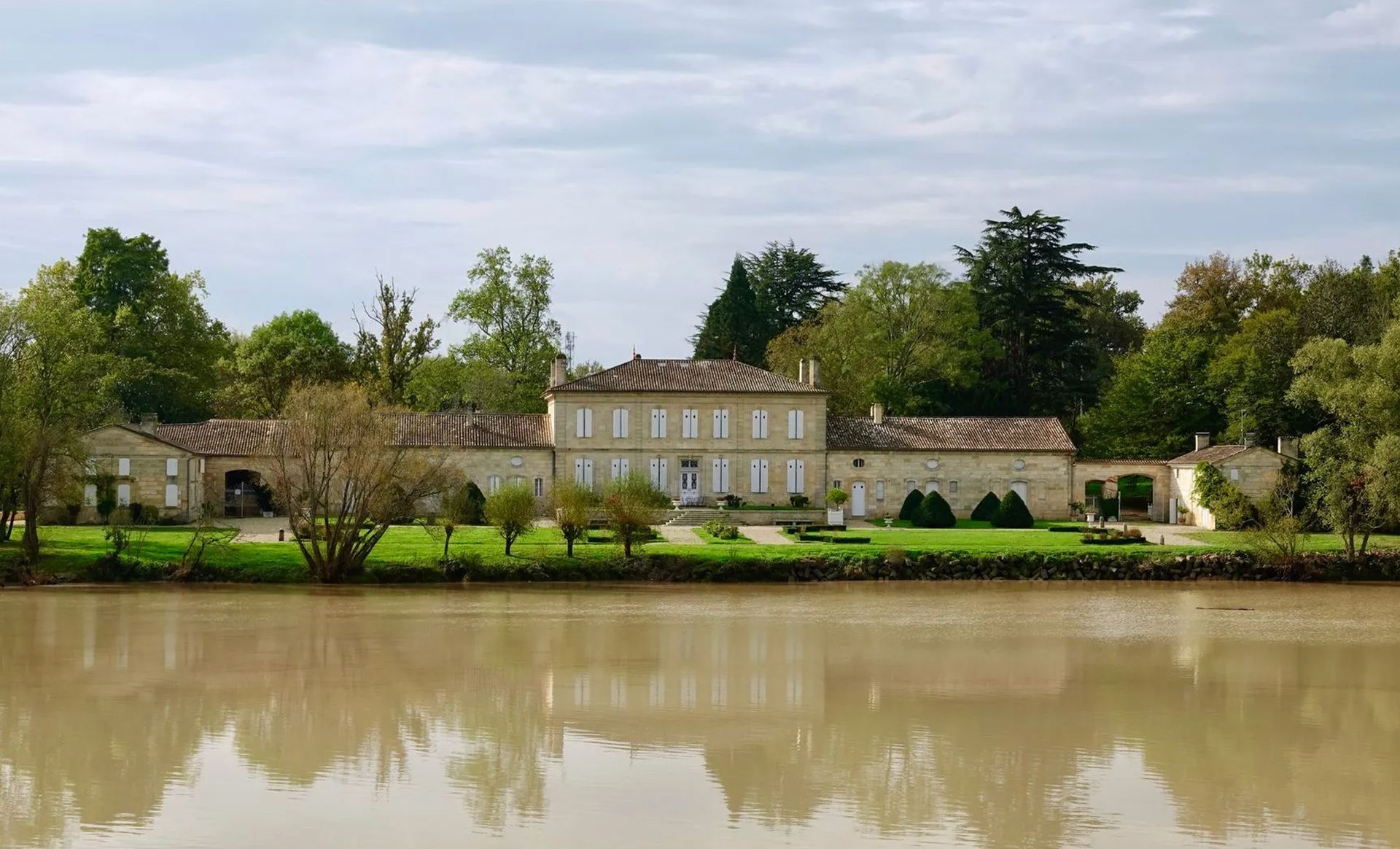 A large house is sitting on the shore of a river surrounded by trees.