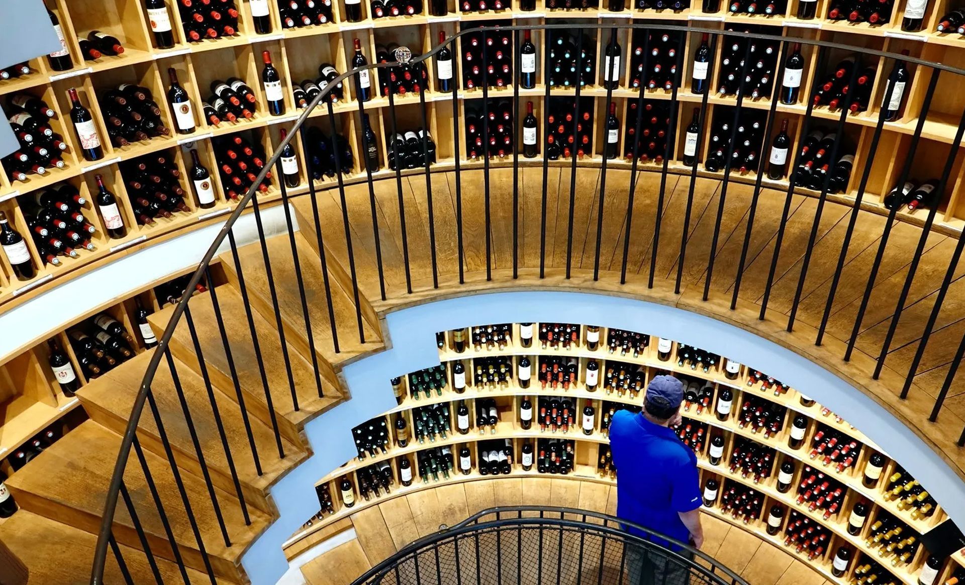 A man in a blue shirt is standing on a spiral staircase in a wine cellar.