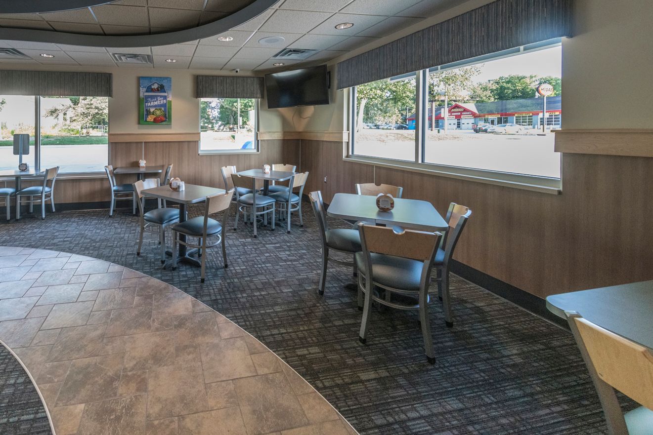Restaurant dining area with tables, chairs, and large windows. Wood paneling, carpeted floor, and a TV.