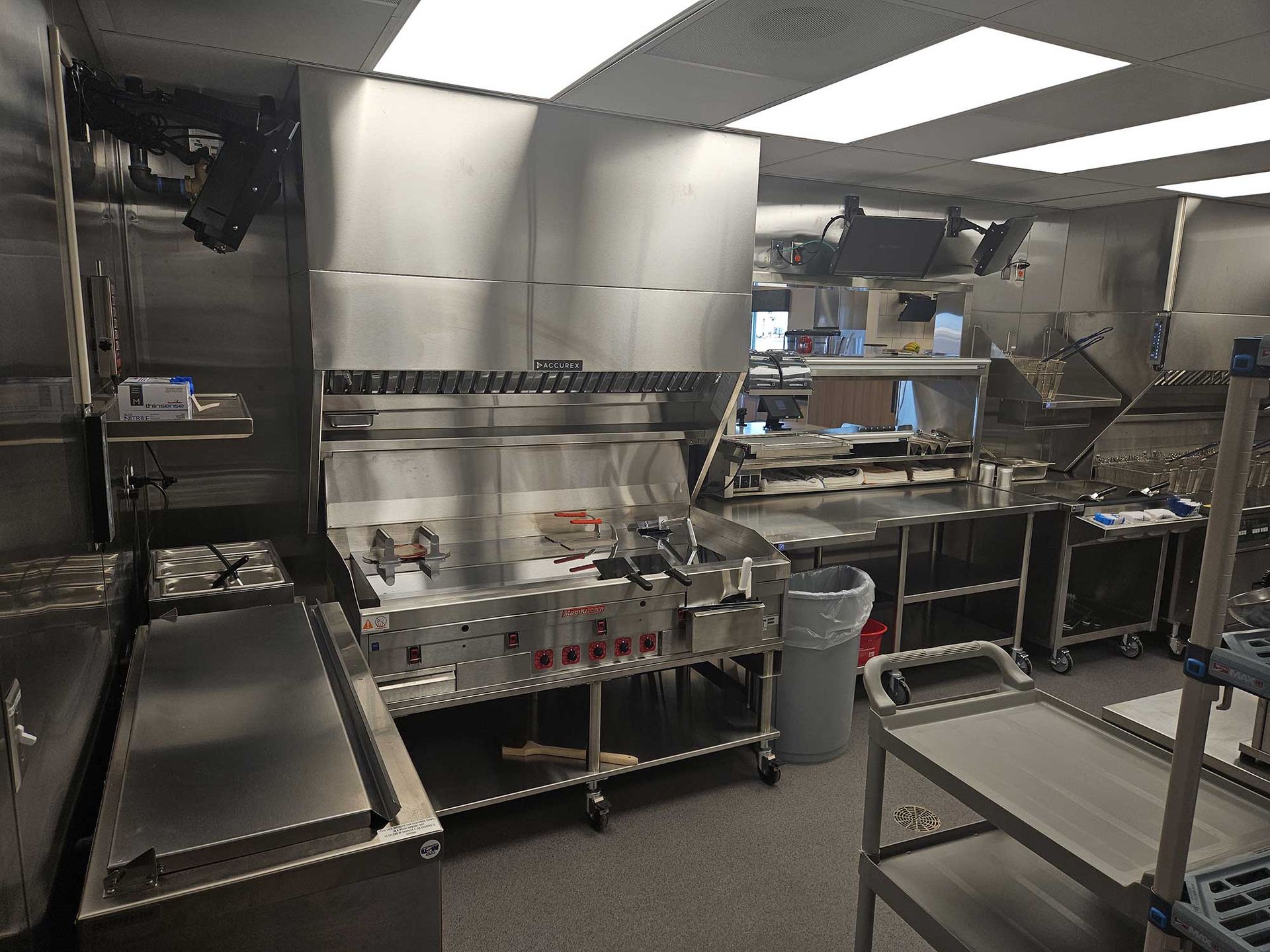 Commercial kitchen with stainless steel appliances, including a grill, hood, and prep tables.